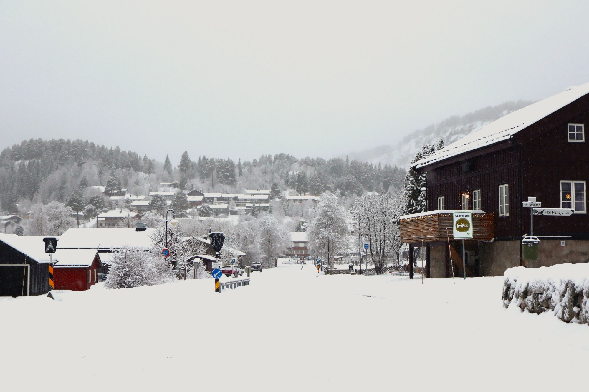 Snødekt vei gjennom et tettsted med hus og trær, omgitt av vinterlandskap og hvite åser i bakgrunnen